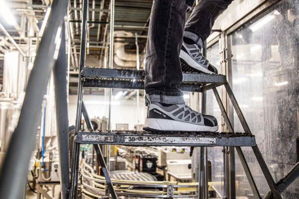 A pair of athletic shoes in black and gray stands on a metal staircase in an industrial space with machines in the background, brightly lit.