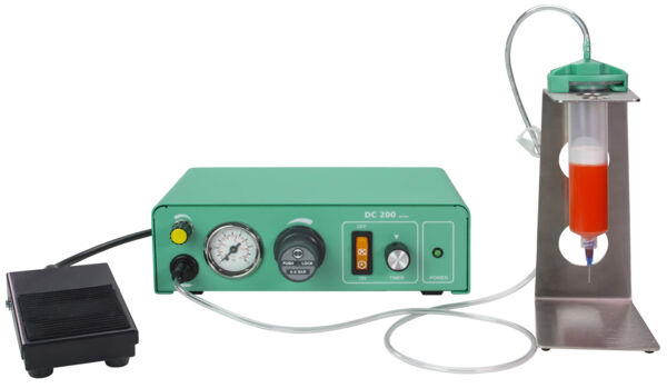 The image shows a green device with a pressure gauge and switches, connected to a transparent container holding a red liquid. A pedal controls the device.