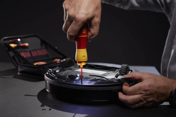 A hand holds a screwdriver and screws on a round, black object, likely a technical device. In the background, a toolbox is visible.