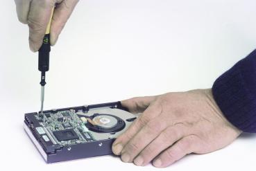 A hand holds a screwdriver, which is inserted into a hard drive. A second hand rests on the hard drive, showing the open circuit board and a fan. The background is neutral.