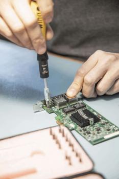 A hand holds a screwdriver and repairs a green circuit board. On the table, more tools are placed in a pink container fitted with slots for various screwdrivers.