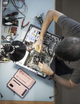 A person is working on an open computer. A component is being mounted with a screwdriver. Tools and accessories are scattered around on a table. A desk lamp provides light.