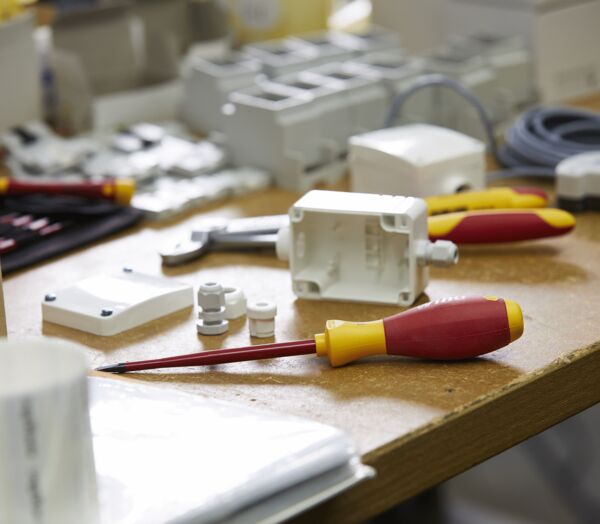 On a table, various tools and components are scattered. There are screwdrivers with red handles, cables, plastic boxes, and screws all over the place. The environment appears crafty and chaotic.