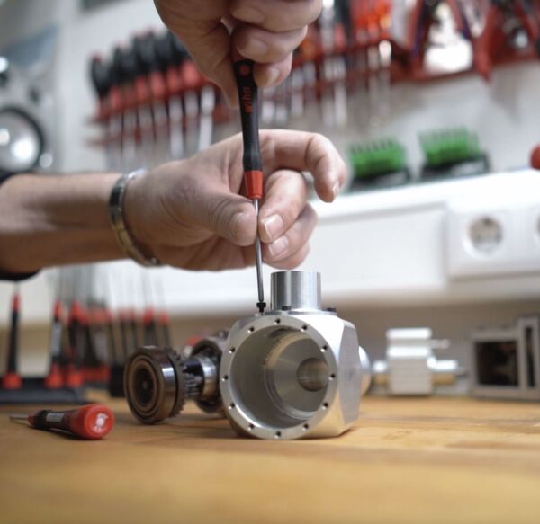 A hand with a screwdriver repairs a metallic part on a wooden table. In the background, various tools and a tool holder are visible.