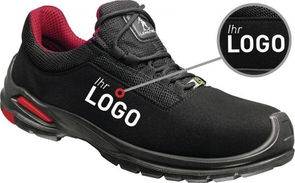 The image shows a black sports or safety shoe with red accents. It has a padded tongue and a non-slip sole. A circular area highlights the logo.