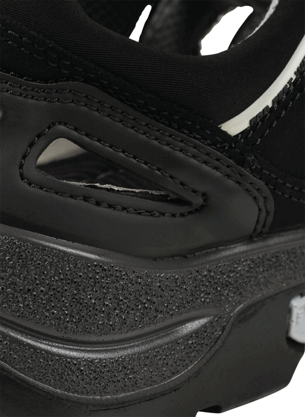 The image shows a part of a black sports shoe. The sole is thick and textured, with smooth and rough areas. There are cutouts on the side that give it a dynamic shape.