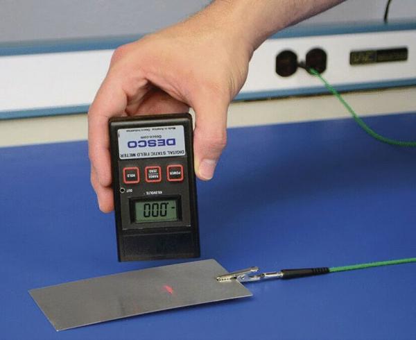 In the picture, a hand is holding a measuring device that displays digital values. In front of the device, there is a flat, rectangular metal plate. A cable with a plug is connected.