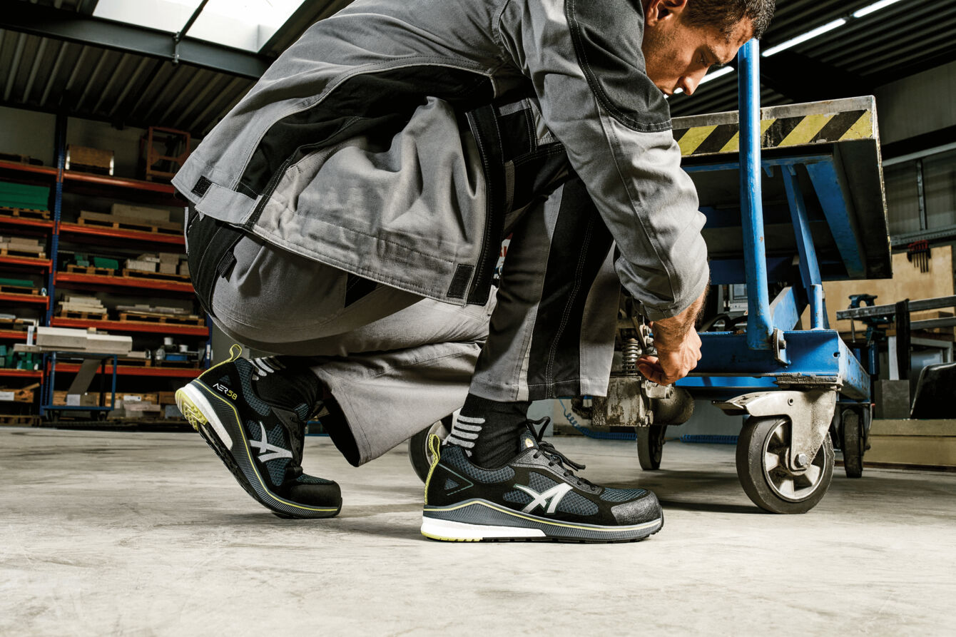 A man is crouching in a room with work materials. He is wearing a gray work suit and black sneakers. His hands are busy as he works on a blue cart.