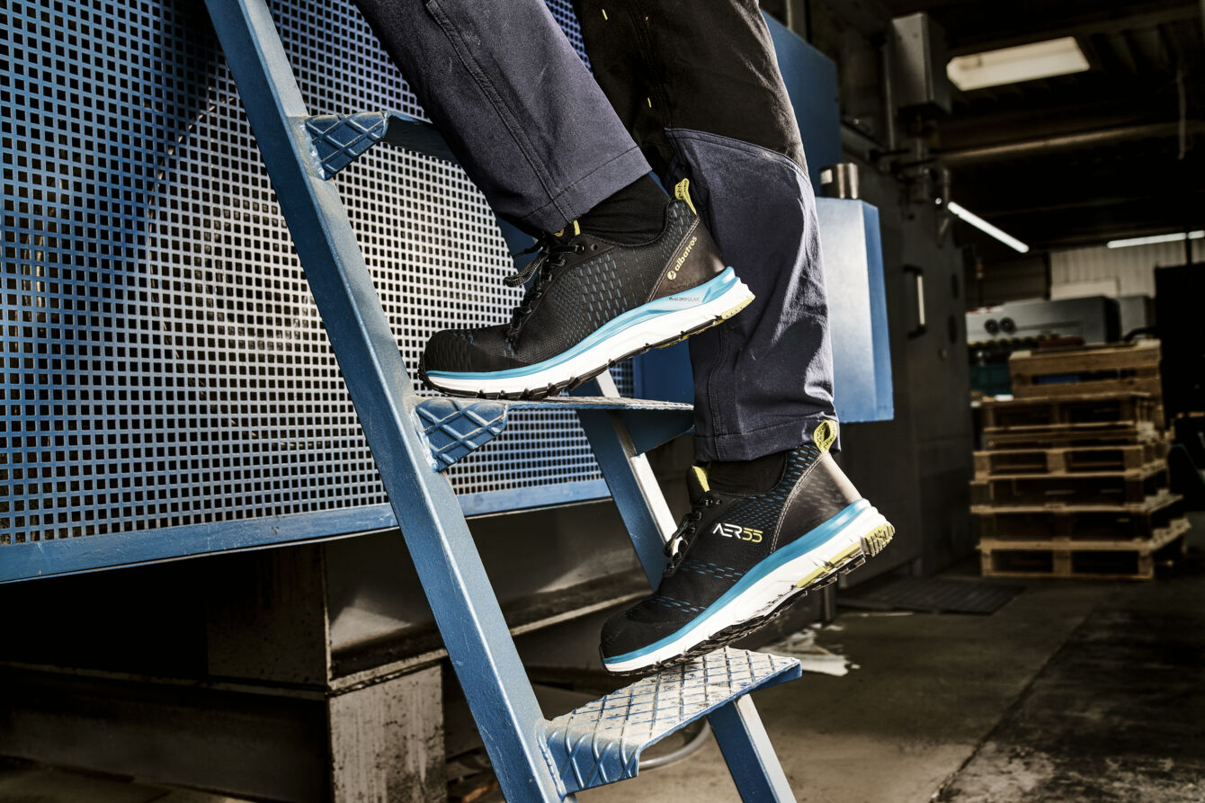 The image shows a person climbing a blue ladder with special work shoes. The shoes are sporty and provide good grip, and the background appears industrial.