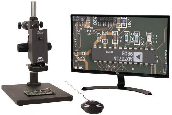 The image shows a microscope on a stable table, next to a monitor displaying a detailed view of a circuit board with electronic components. A mouse is also visible.