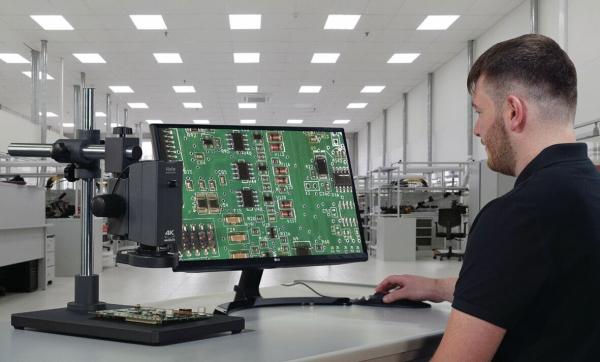 A man is sitting at a table, looking at a green circuit board with components on a monitor. The room is bright and modern, with additional workstations in the background.