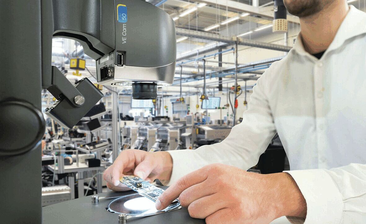 A person in a white shirt is working at a table, examining a circuit board under a microscope. In the background, machines can be seen in a bright factory.