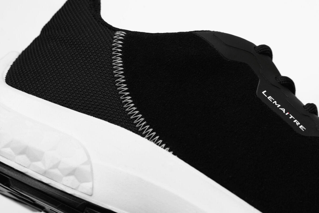 The image shows a sporty shoe, predominantly in black with a white sole. The shoe has a smooth surface and striking, bright stitches on the side.