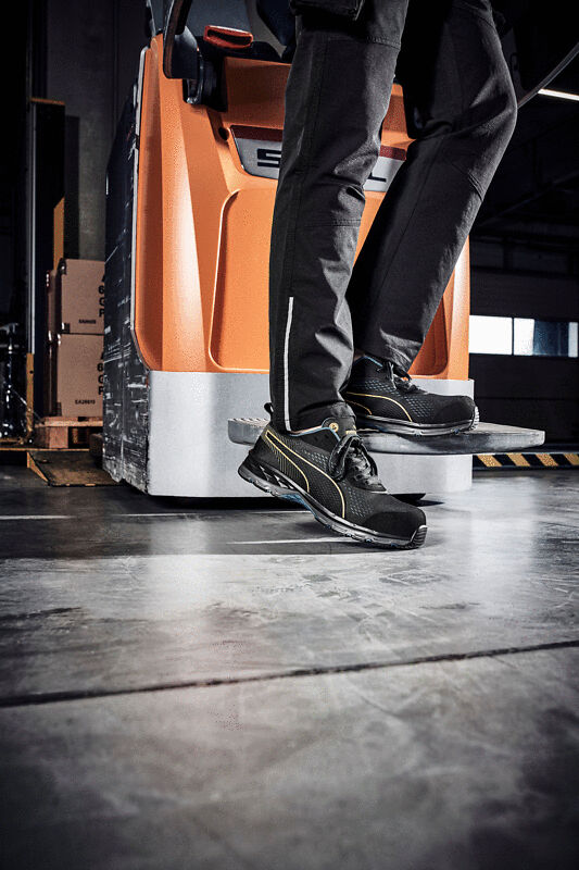 The image shows a worker standing on a platform with safety shoes. In the background, there is an orange device placed on a smooth floor.