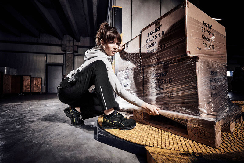 A person in sportswear is squatting next to stacked boxes wrapped in stretch film. The floor is gray, and the background shows a warehouse with dim lighting.