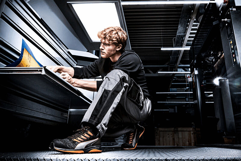 A young man in work clothes is sitting on a metal floor studying documents. He has curly, light hair and is wearing shoes with orange accents. Behind him is an industrial space.