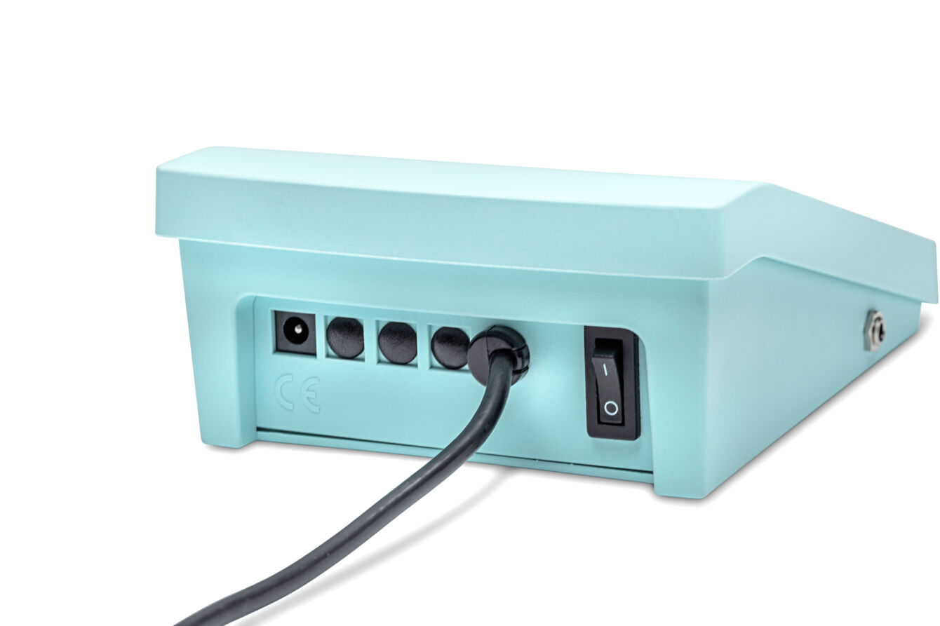 The image shows the back of a turquoise-colored device. It has multiple ports for cables and a power switch. A black cable is plugged in. The shape is rectangular and flat.