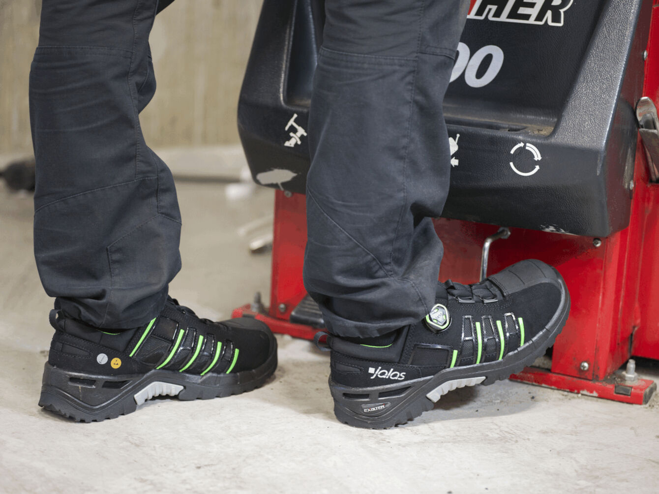 The image shows feet in black work shoes with green accents. A person is standing next to a machine unit in an industrial setting. The floor is bright and smooth.