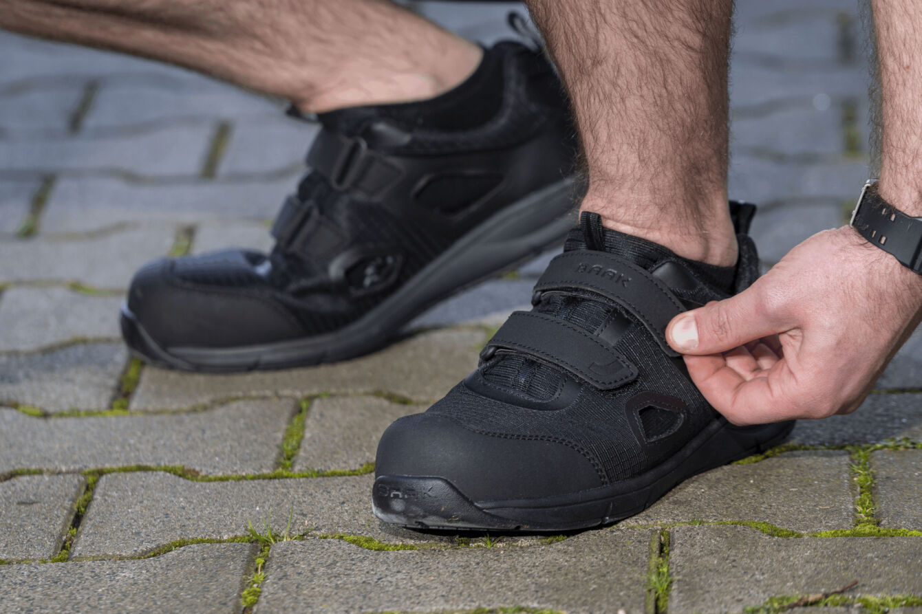 The image shows a man tightening a black Velcro shoe with one hand. He is standing on a cobblestone path with green moss between the stones.