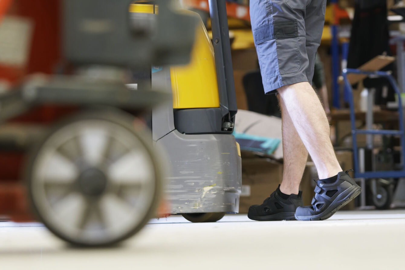 The image shows the legs of a person in shorts and black shoes. In the foreground, there is a wheel, likely from a cart or piece of equipment. The background is blurry.