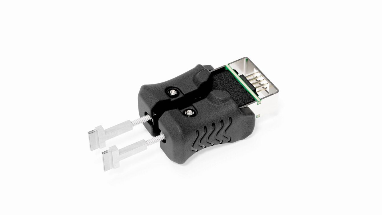 Das Bild zeigt einen schwarzen Adapter mit zwei silbernen Stiften, die nach vorne ragen. Oben ist ein Anschluss sichtbar, auf dem eine grüne und schwarze Fläche zu sehen sind.