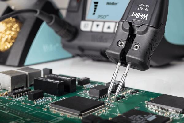 The image shows a circuit board with several black and silver chips. A tool that looks like a pair of pliers is holding one of the chips. In the background, you can see a soldering iron device.