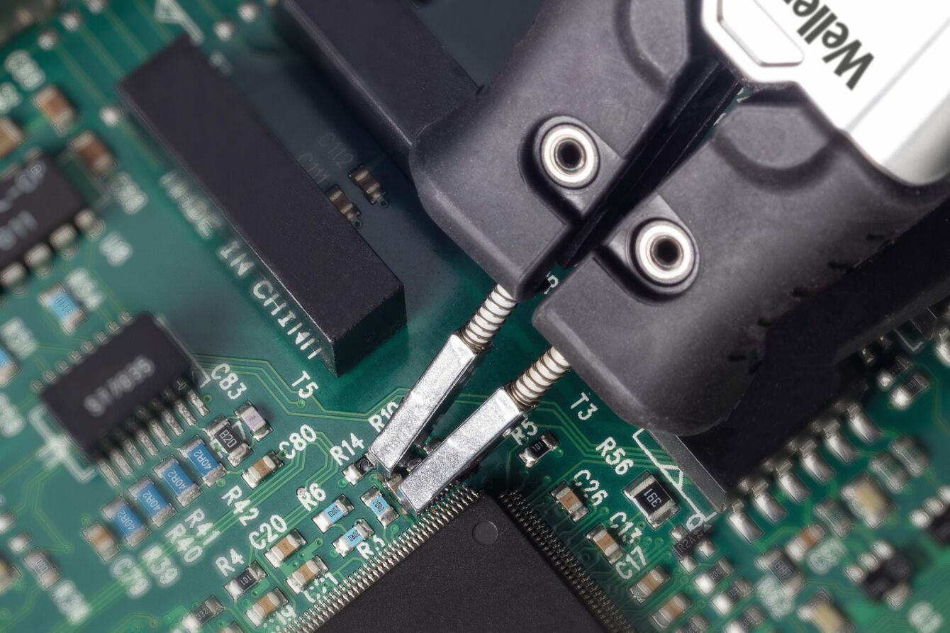 The image shows a green circuit board with electronic components. Two pliers precisely grip a spot on the board. Detailed circuits are visible.