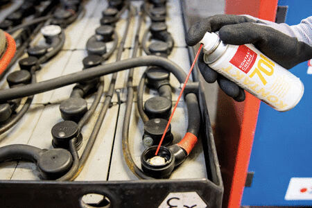 A hand wearing a black glove is spraying a product from a can onto the contacts of a large, rectangular battery with many wires. The background is blue.