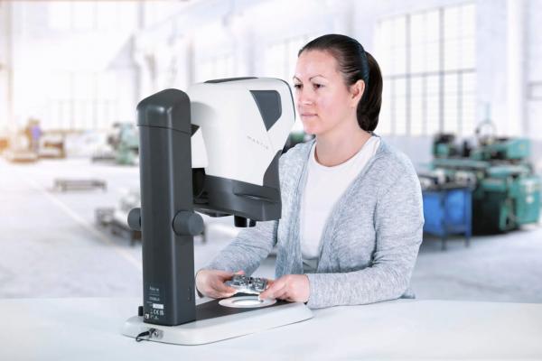 A woman is sitting at a table, in front of her there is a modern device with a monitor. She is focused on it, while in the background a bright workshop is visible.