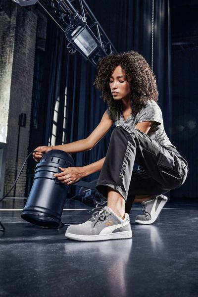 A woman with curly hair is kneeling on a dark floor. She is wearing gray sweatpants and shoes. In one hand, she holds a black kettlebell, while she looks on intently.