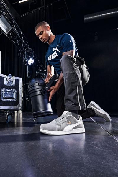 A man in a dark blue T-shirt with the Puma logo bends down and picks up a black lamp. He is wearing gray sneakers and dark, loose pants. The background is modern and industrial.