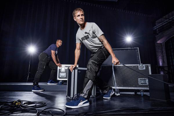 Zwei Männer arbeiten in einer dunklen Umgebung. Einer trägt eine kurze Sporthose und ein T-Shirt, während er ein großes Case hält. Der andere Mann hebt ebenfalls ein Case, steht im Hintergrund.