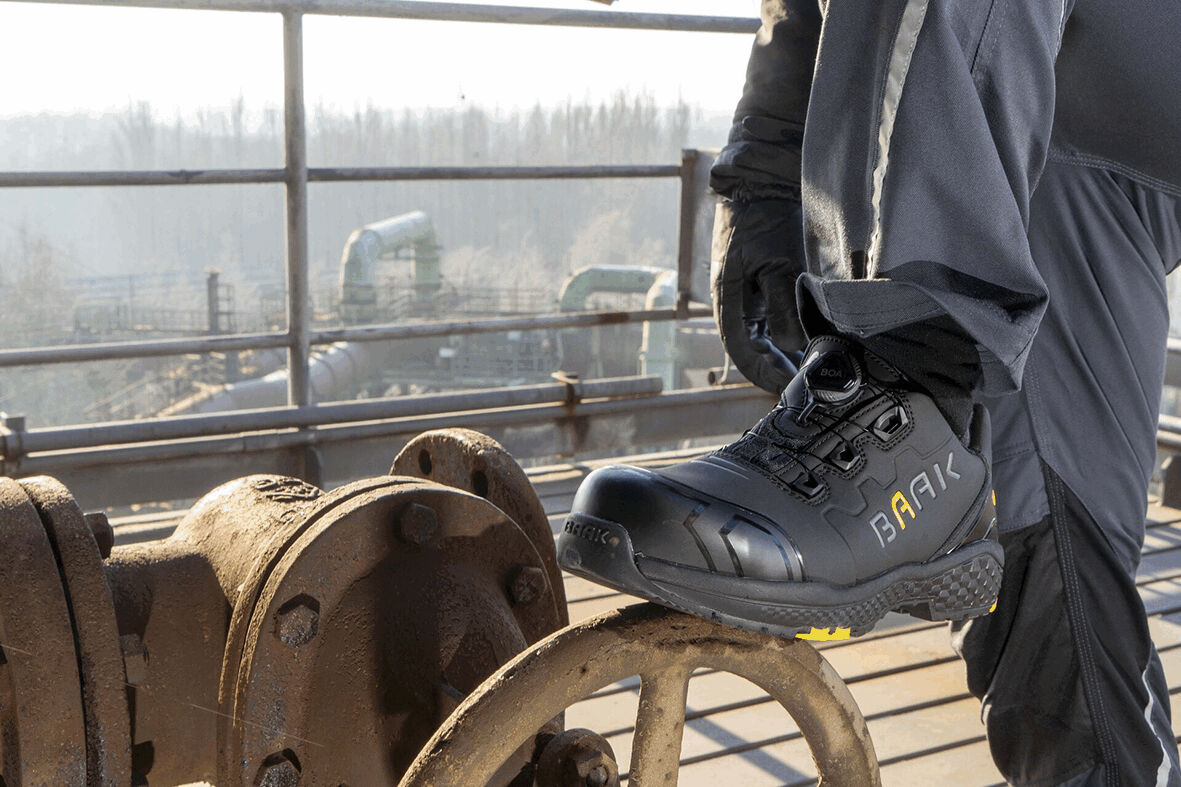 The image shows a foot in a black safety shoe standing on a metallic, round device. In the background, an industrial environment with pipes can be seen.