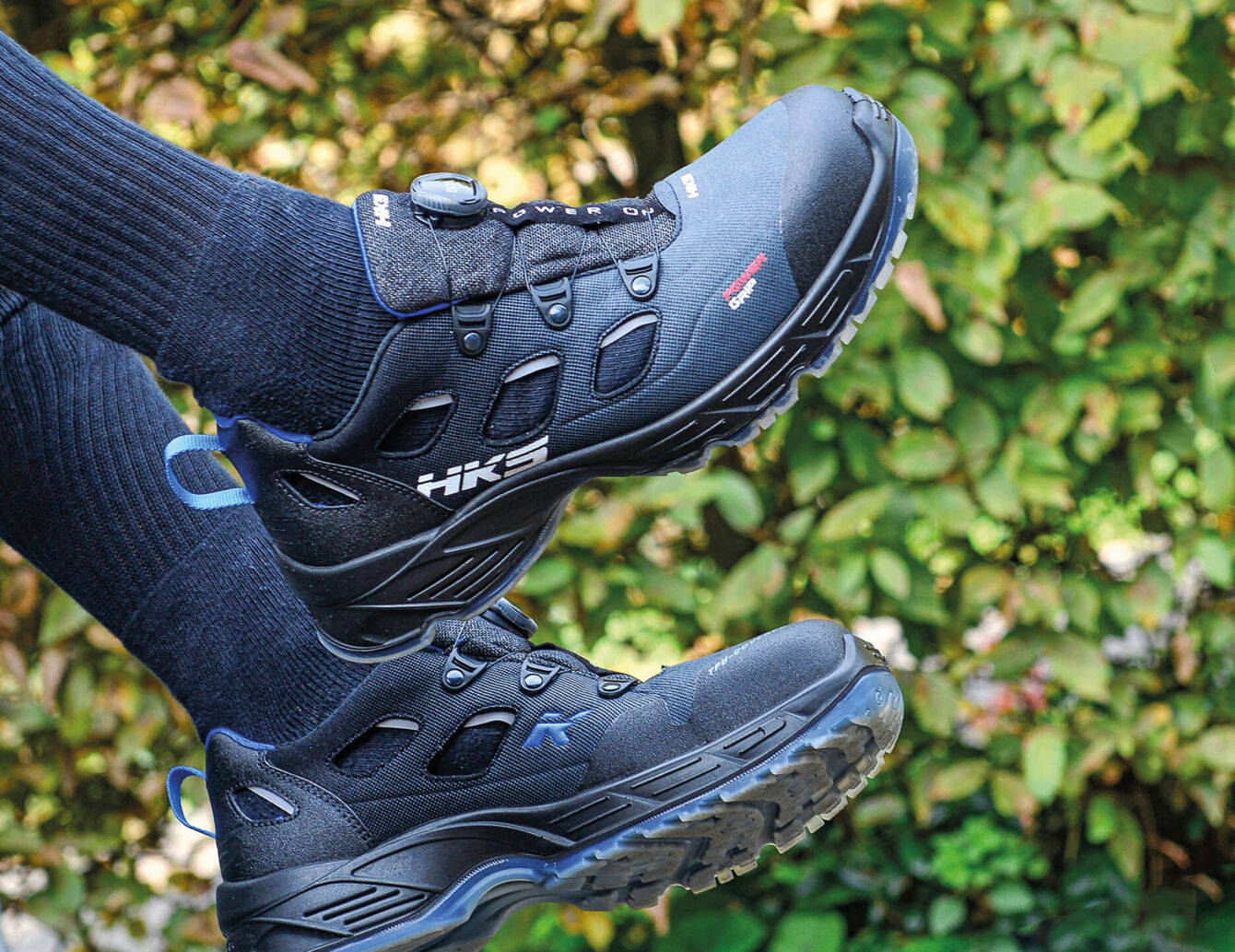 The picture shows a pair of black hiking boots with a sporty design. A person is wearing black socks, and in the background, there are green, blurred plants.