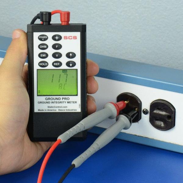 A handheld grounding tester is being held. It displays a number (117) on a green screen. Two red and gray cables are connected to an outlet.