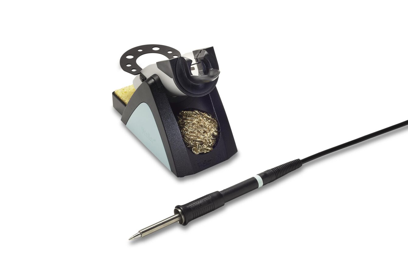 The image shows a soldering station with a semi-circular stand for a soldering iron. It is placed next to a cleaning spiral and a sponge for cleaning the soldering tip.