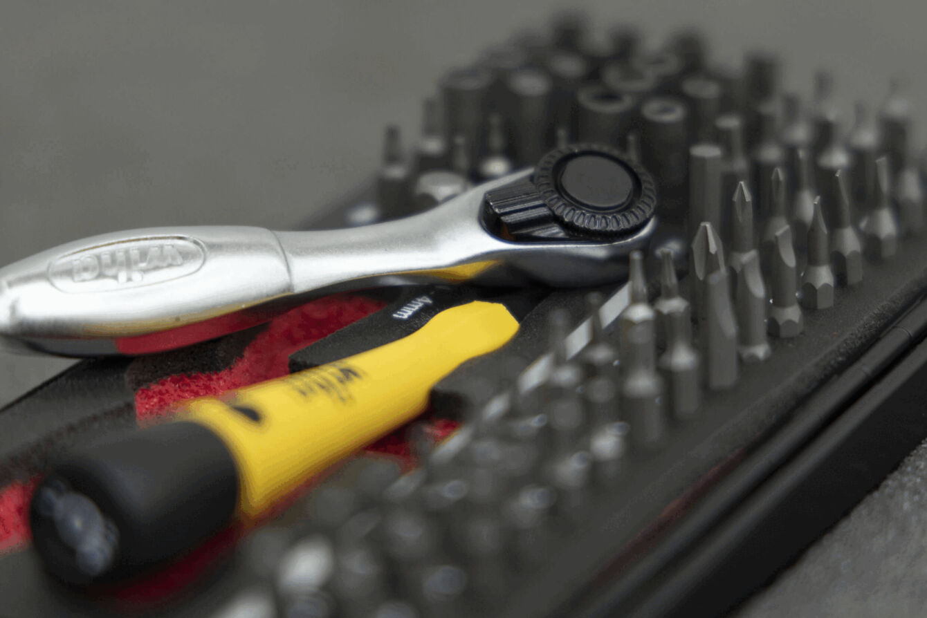 The image shows various tools. A silver ratchet lies next to a yellow screwdriver, surrounded by many black bits arranged in a red holder.