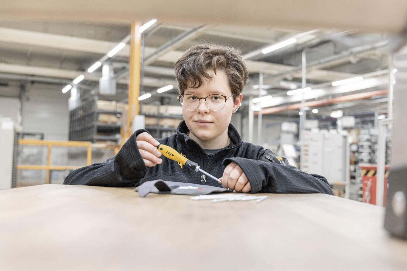 Ein Junge mit kurzen, braunen Haaren und einer Brille sitzt an einem Tisch in einer Werkstatt. Er hält ein Werkzeug in der Hand und schaut konzentriert auf eine graue Materialprobe.