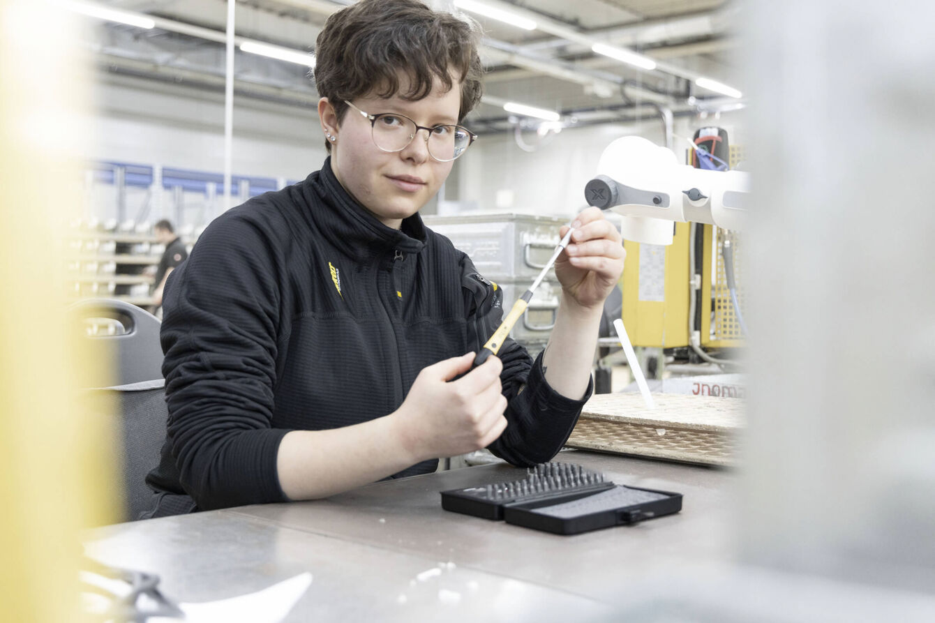 Eine Person mit kurzem, lockigem Haar sitzt an einem Tisch in einer Werkstatt. Sie trägt eine schwarze Jacke und hält ein Werkzeug in der Hand, während sie konzentriert in die Kamera schaut.