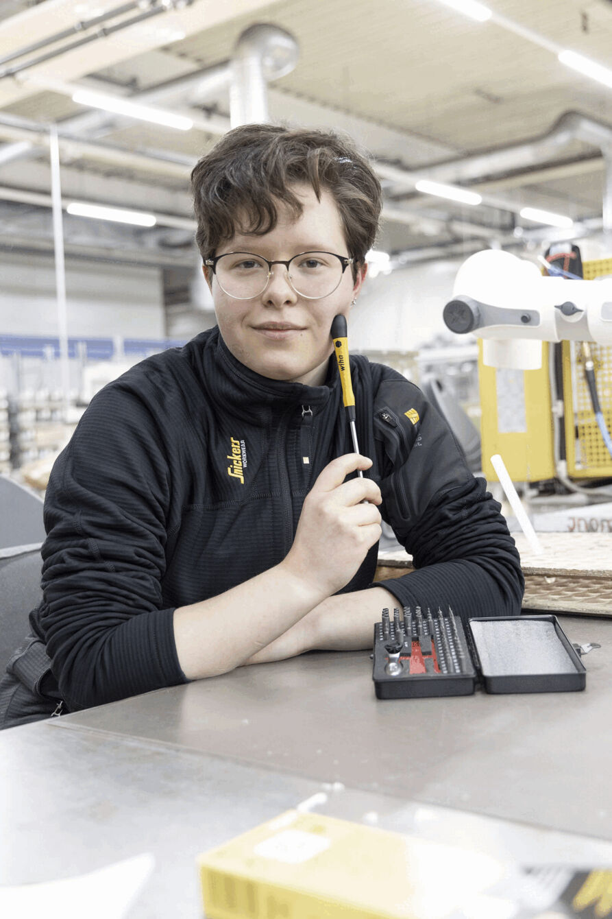 Eine Person mit lockigem, braunem Haar und Brille sitzt an einem Tisch. Sie hält einen Bleistift in der Hand, vor ihr liegt ein Werkzeugkasten mit verschiedenen Schraubendrehern.