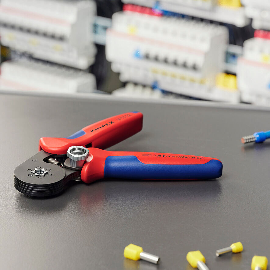 The image shows a crimping tool with red and blue handles on a dark table. In the background, colored terminal connectors and cables are visible.