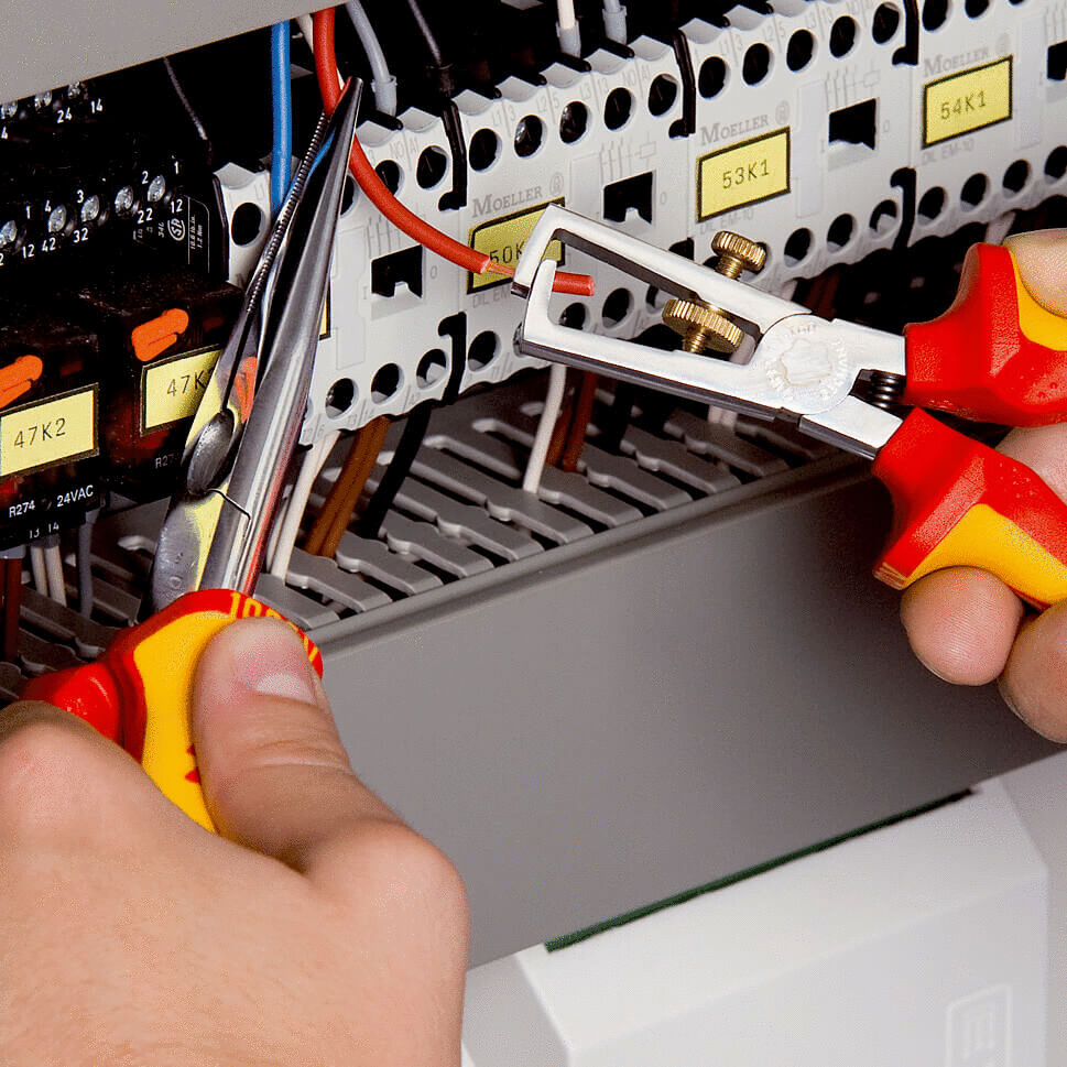 Two hands hold tools. One pair of pliers grasps a wire, while the other is about to cut a wire. In the background, electrical components with labels are visible.