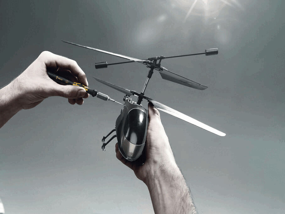 A person is holding a small model helicopter and is screwing on one of the rotors with a screwdriver. The background is bright and neutral.