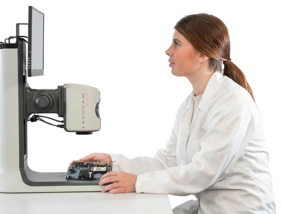 A woman in a white lab coat is sitting at a table. In front of her, there is a screen and a microscope. She is holding a circuit board in her hand and looks focused.