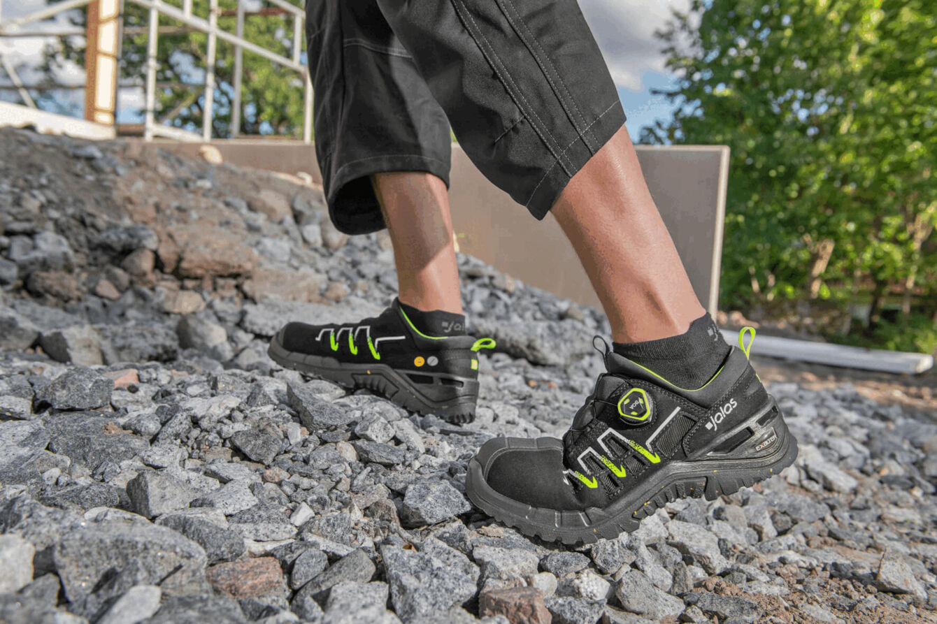 The image shows a foot in a black, sporty shoe with green accents. The shoe is on uneven, rocky ground. In the background, trees and a construction site are visible.