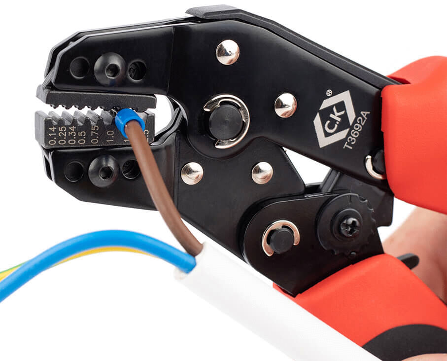 The image shows a crimping tool with red handles. In the tool, there are three wire leads: a green, a blue, and a brown one. The tool has a precise opening for crimping.