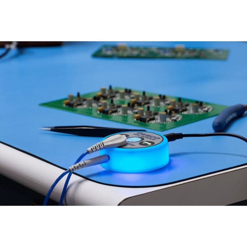 On a blue table lies a glowing, round adapter with cables. In the background, two green circuit boards are visible, equipped with various electronic components.