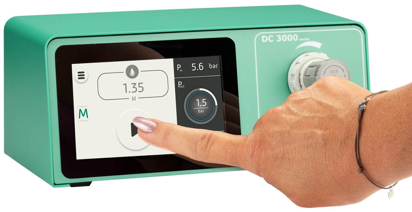 The image shows a green device with a touchscreen. A hand is touching the screen. On the display, numbers and symbols are visible that indicate various settings.