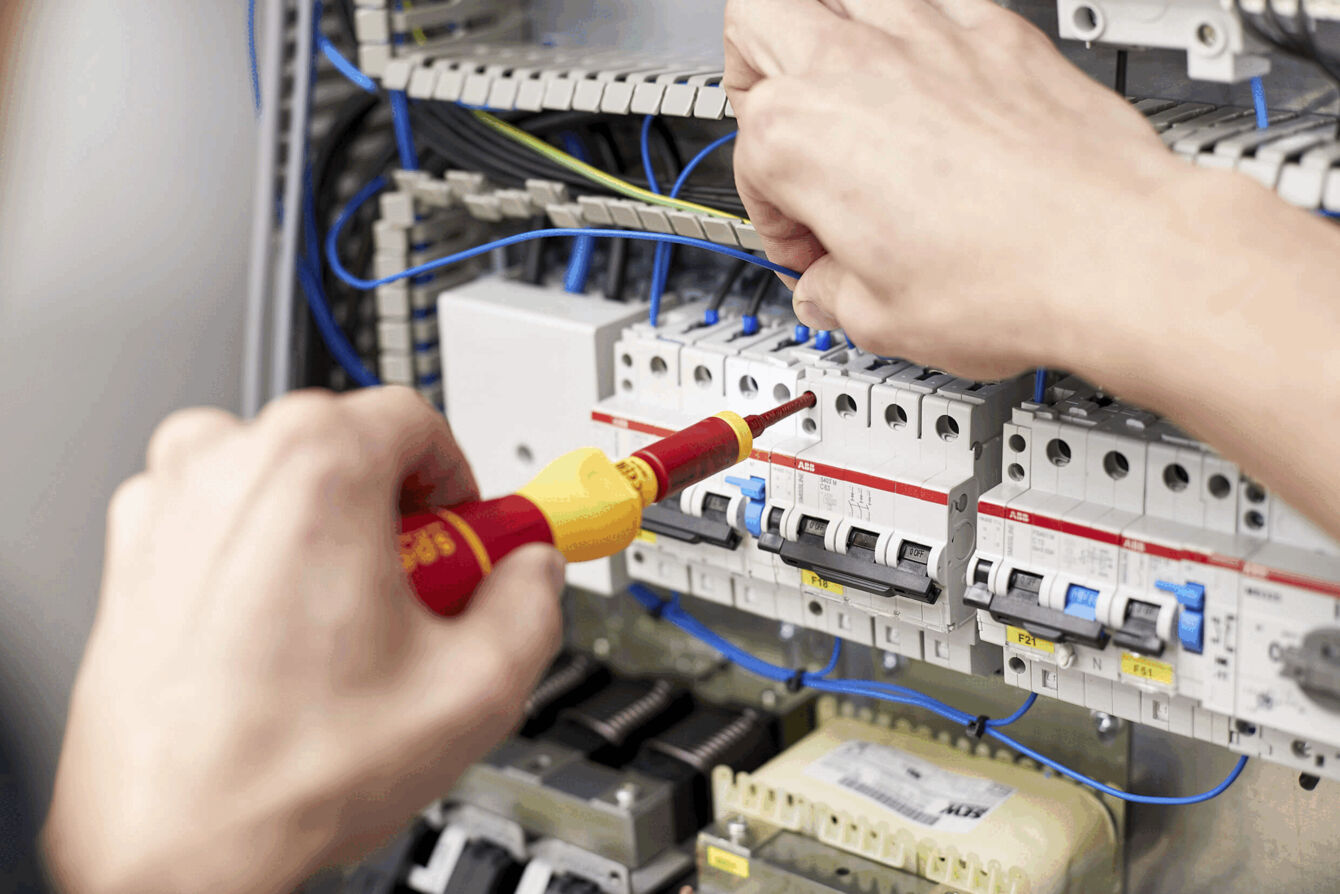 A person is working on a control cabinet with electrical components. In one hand, they are holding a screwdriver. Several fuses and cables are visible. A technical environment.