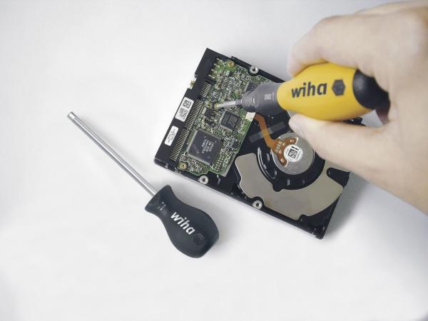 The image shows a hand pointing a screwdriver at the circuit board of a hard drive. Next to it lies another screwdriver with a black handle. The background is light.